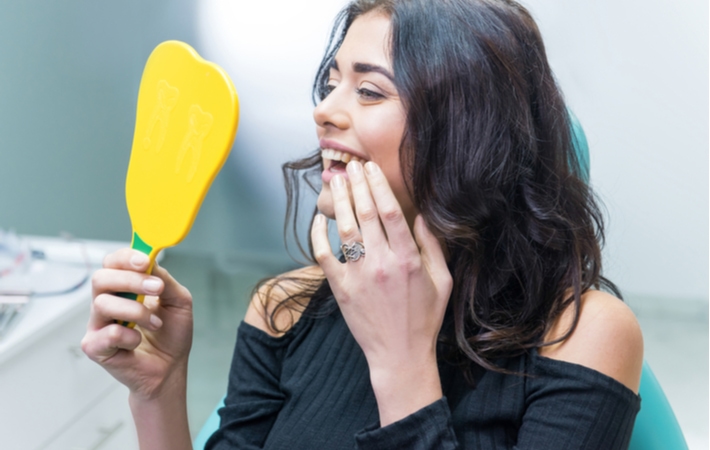 A woman in a black shirt at the dentist using a yellow handheld mirror to look at her new, placed dental implants