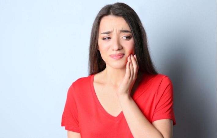 A woman wearing a bright red shirt, using her left had to hold her cheek while she winces in pain from a broken tooth