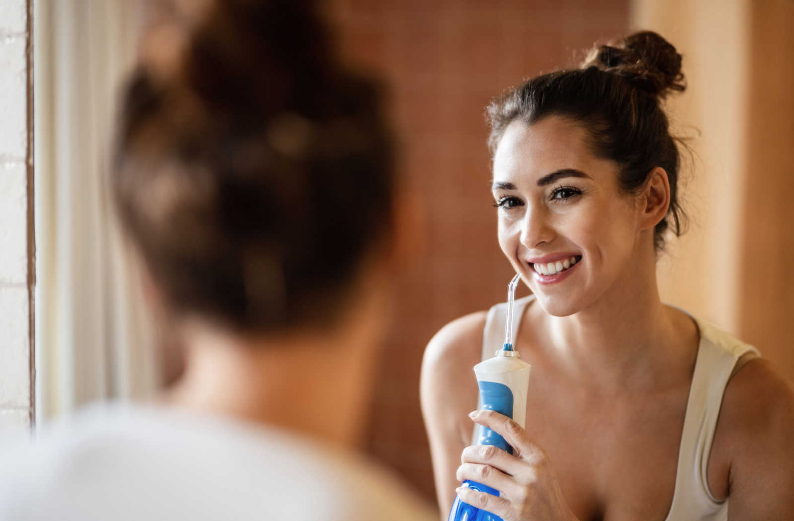 A woman looking in the mirror while using a water flosser to help maintain good oral hygiene.
