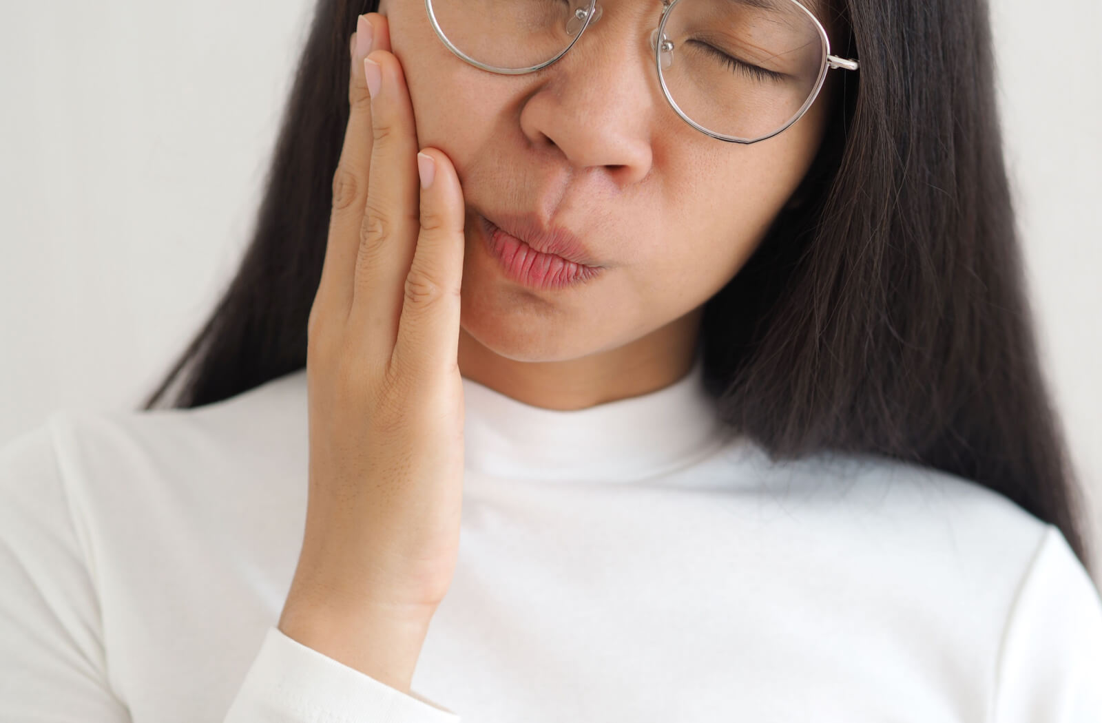 A lady in a white longsleeve with her eye glasses on is holding her right jaw in her right hand with a gesture of pain on her face.