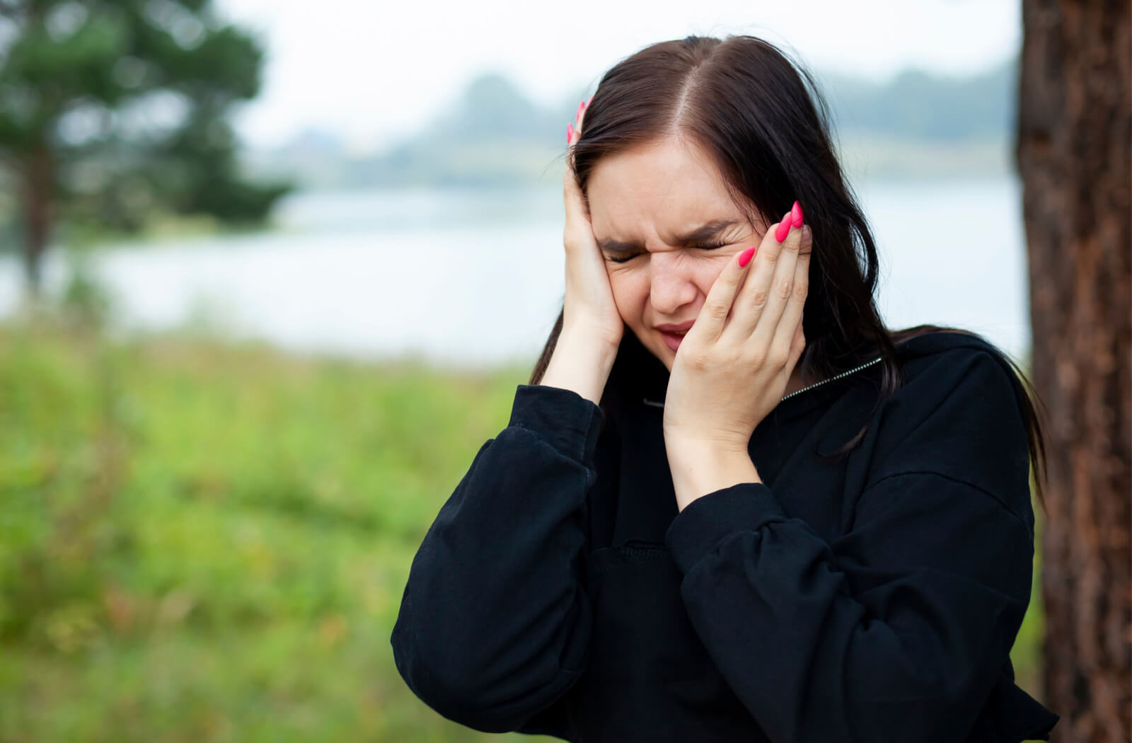 A young woman outdoors holding her head and jaw and squinting due to jaw pain and a headache.