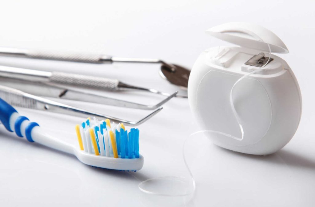 A toothbrush, floss, scaler, and other dental tools which are all important for maintaining good oral health laying on a white surface