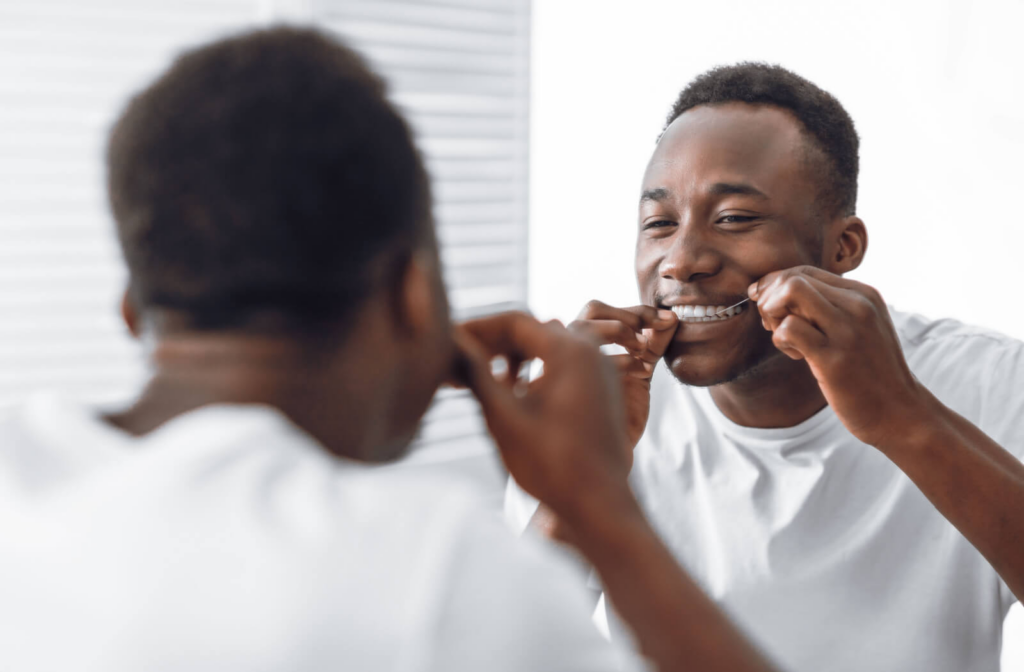 A man flossing his pearly white teeth in front of a mirror.