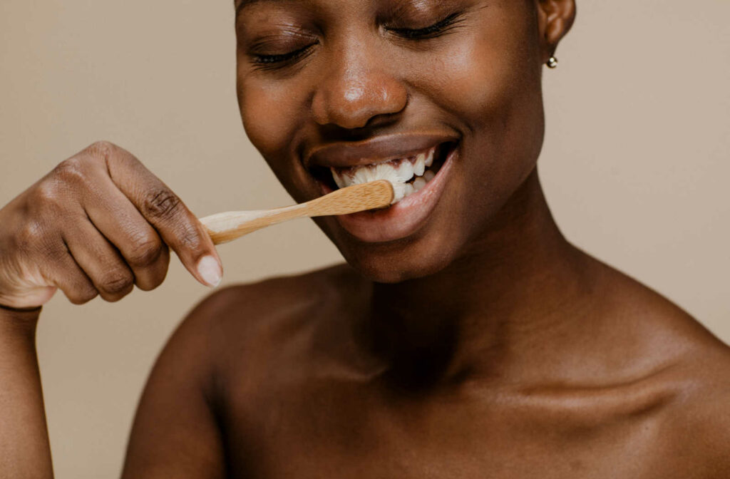 A young woman brushing her pearly white teeth using a wooden toothbrush against a beige background.