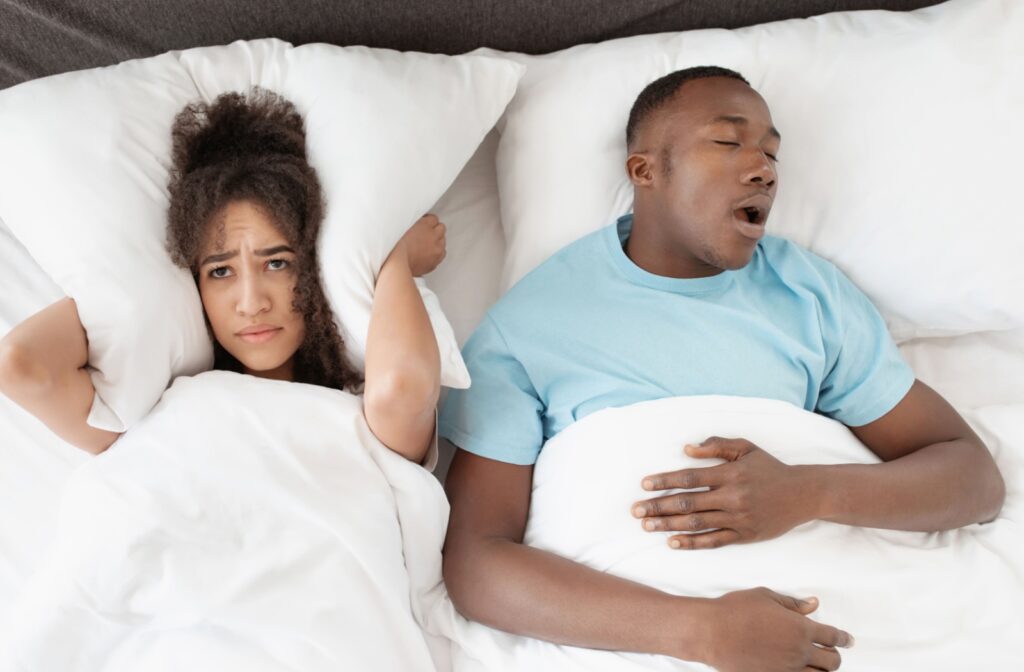 A man and woman in bed, the man is asleep and snoring, and the woman is awake and holding her pillow over her ears.