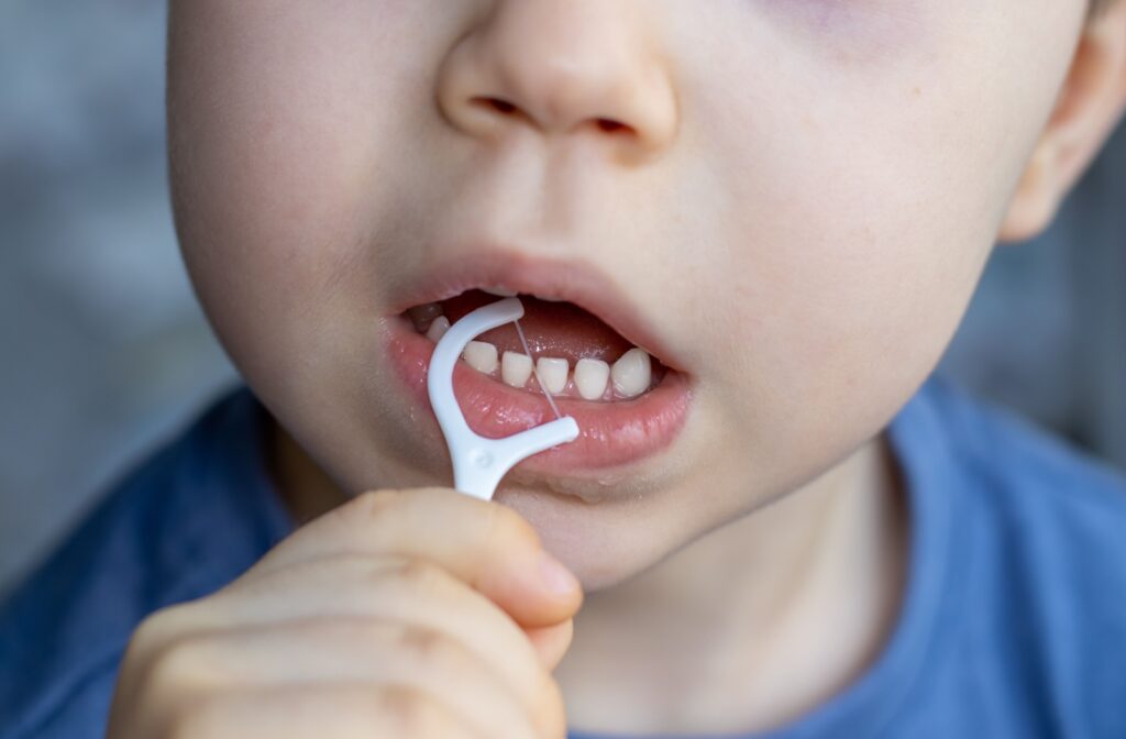 A young child in a blue shirt with gaps in their teeth uses a flossing pick to clean between their teeth