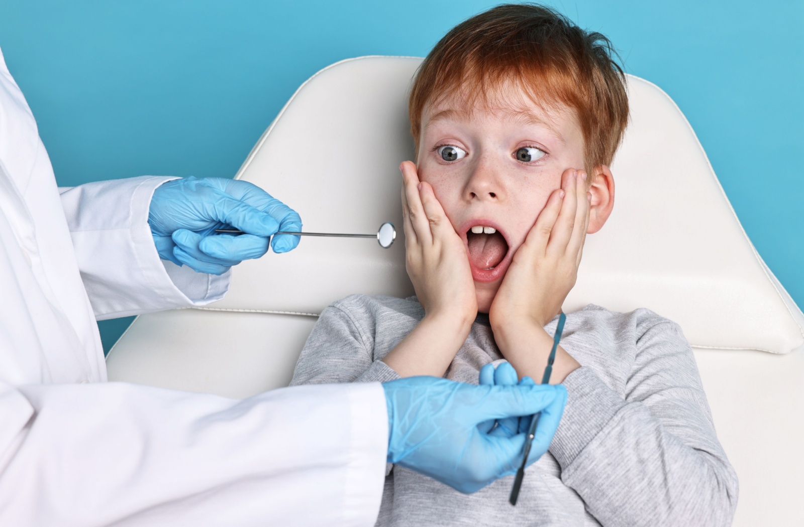 Against a blue background, a young child gasps in fear as a dental hygienist approaches with dental tools