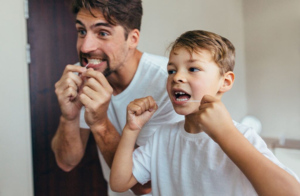 An adult and a child floss their teeth together in front of a mirror.